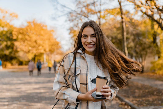 A woman with long light brown hair smiling in an autumn park while holding sunglasses, a cup of coffee, and a phone.