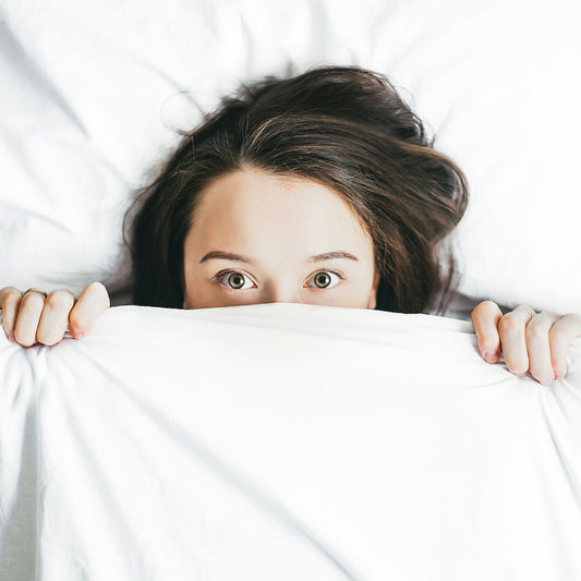 Woman with brown hair in bed under covers, looking at camera.
