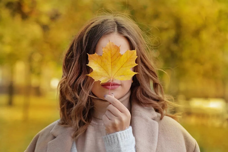 A woman with honey blonde hair holds a bright yellow maple leaf in front of her face.