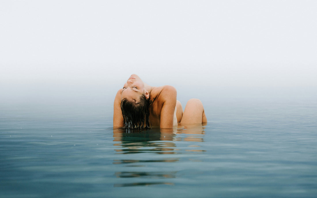 Woman emerging from calm water with wet hair slicked back, highlighting how swimming impacts hair extensions in a serene, misty setting.
