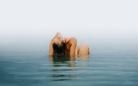 Woman emerging from calm water with wet hair slicked back, highlighting how swimming impacts hair extensions in a serene, misty setting.