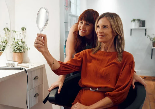 An older woman admires herself in the mirror after trying a hair topper with the stylist at her shoulder.