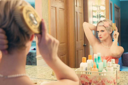 A young woman wearing a wig, brushing her hair in the mirror.