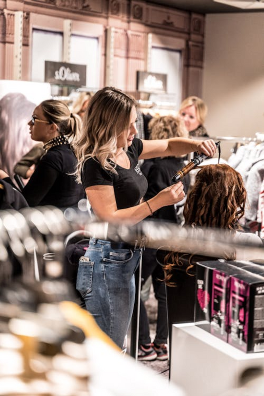 A hairstylist using a curling iron to style a client's hair extensions in a busy salon, with other stylists and customers visible in the background. Source: Pexels.