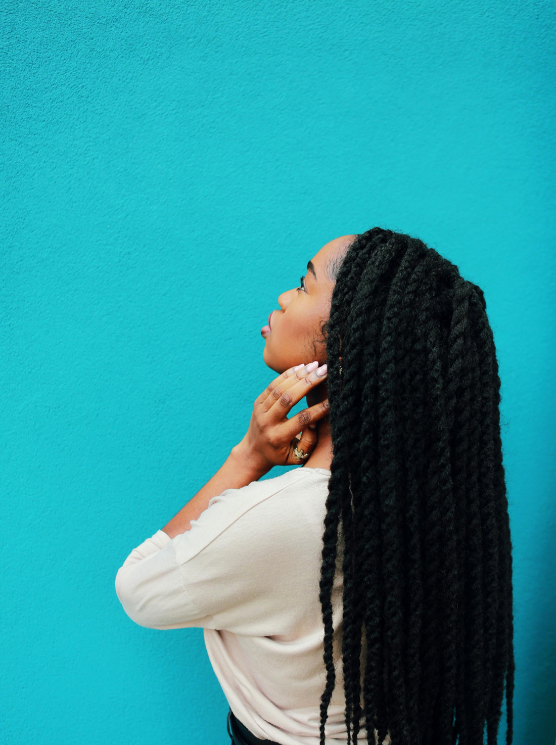 A woman with dark skin and long, thick twist braids looks upwards thoughtfully. She wears a white blouse and stands against a vibrant turquoise blue wall. Her hands gently touch her neck, adding a touch of elegance to her thoughtful pose.