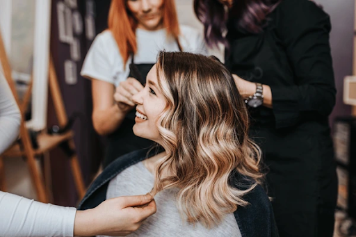  Smiling woman at a salon with freshly styled wavy hair, while hairstylists adjust and finish her look.