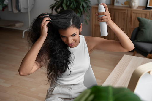 Image of a lady with dark long hair spraying dry shampoo into her roots.