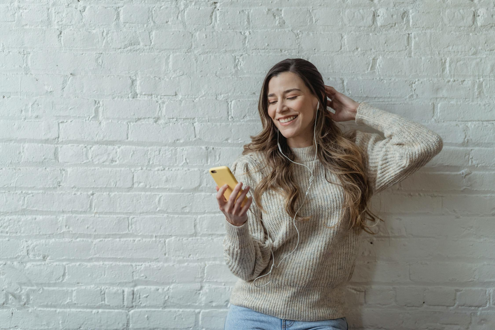 A woman with wavy hair on a video call against a white brick wall.