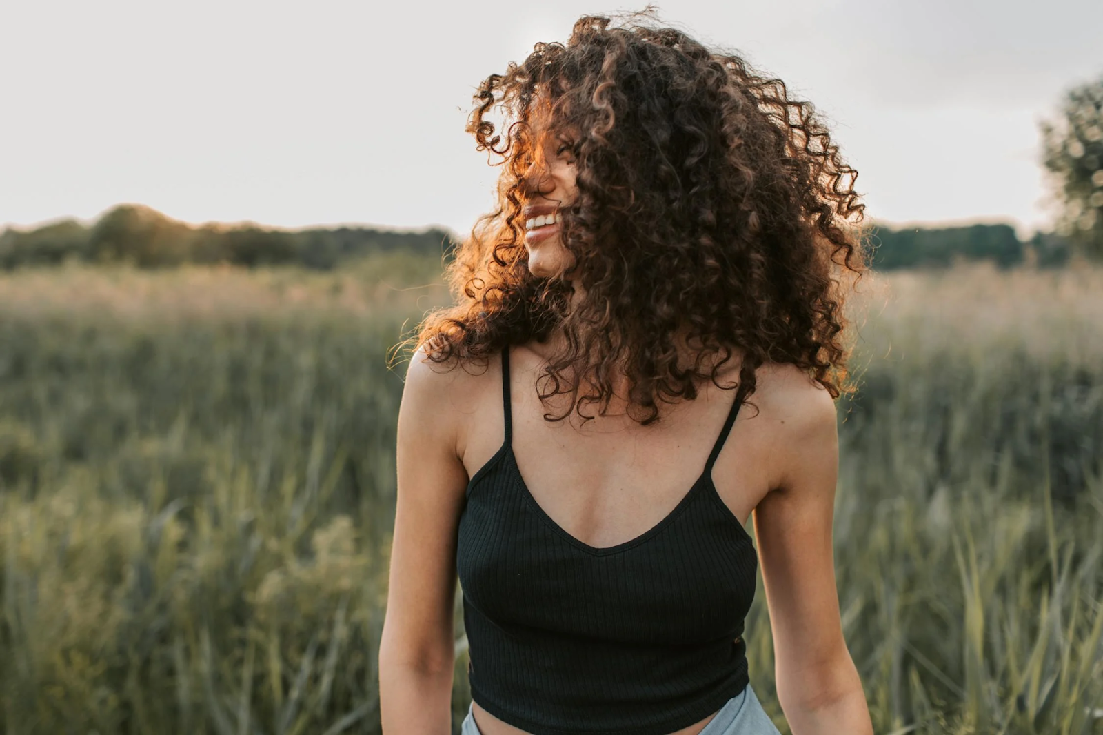 A young smiling woman with a curly hair type, walking through a field.