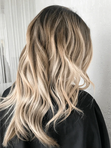 Back view of long, wavy hair featuring a beige blonde balayage with dark roots and soft, blended highlights.