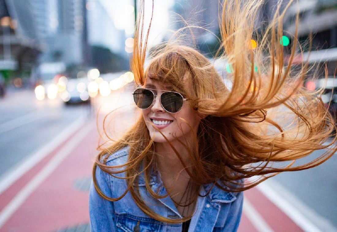 A smiling young woman wearing aviators with copper hair blowing in the wind.