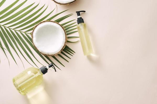 Flat lay of natural hair care ingredients featuring halved coconuts, glass pump bottles filled with golden oil, and a green palm leaf on a neutral beige background.