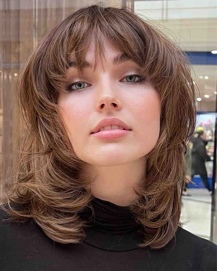 Woman with a layered, medium-length shag haircut featuring soft feathered ends and curtain bangs, wearing a black high-neck top