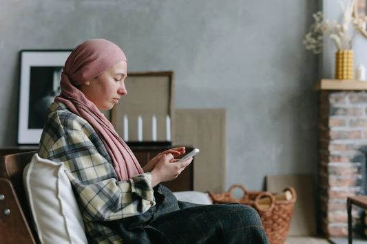 A woman with hair loss wearing a headscarf, sitting on the sofa and scrolling on her phone.