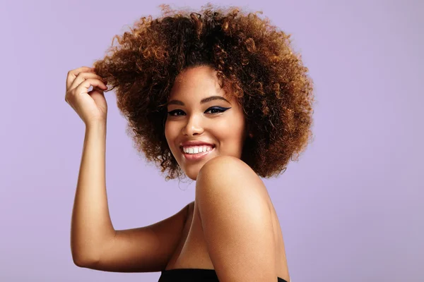 Smiling woman with voluminous curly hair posing against a pastel purple background. She wears bold eyeliner and a strapless black top, playfully lifting a curl with her hand.