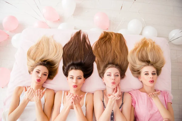 Four young women with different hair colors lying on a bed with their hair fanned out, blowing kisses towards the camera. The setting includes soft pink and white balloons, creating a playful and celebratory atmosphere. 