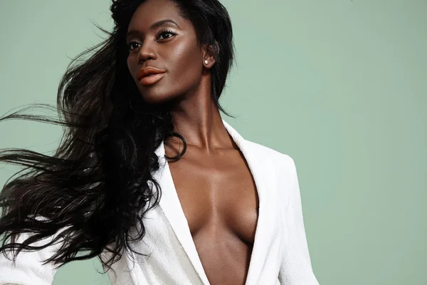 Elegant Black woman with long, flowing black hair wearing a stylish white blazer against a muted green background. She gazes to the side with a poised expression and confident look, her hair captured in motion. 