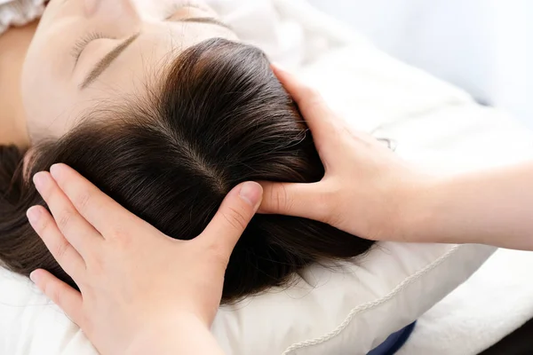 Close-up of a woman’s head receiving a relaxing scalp massage, with hands gently pressing on both sides of the head while lying on a pillow.