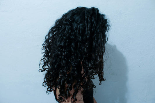 Woman with long, dark, curly hair standing sideways against a light blue wall, her face turned away from the camera.
